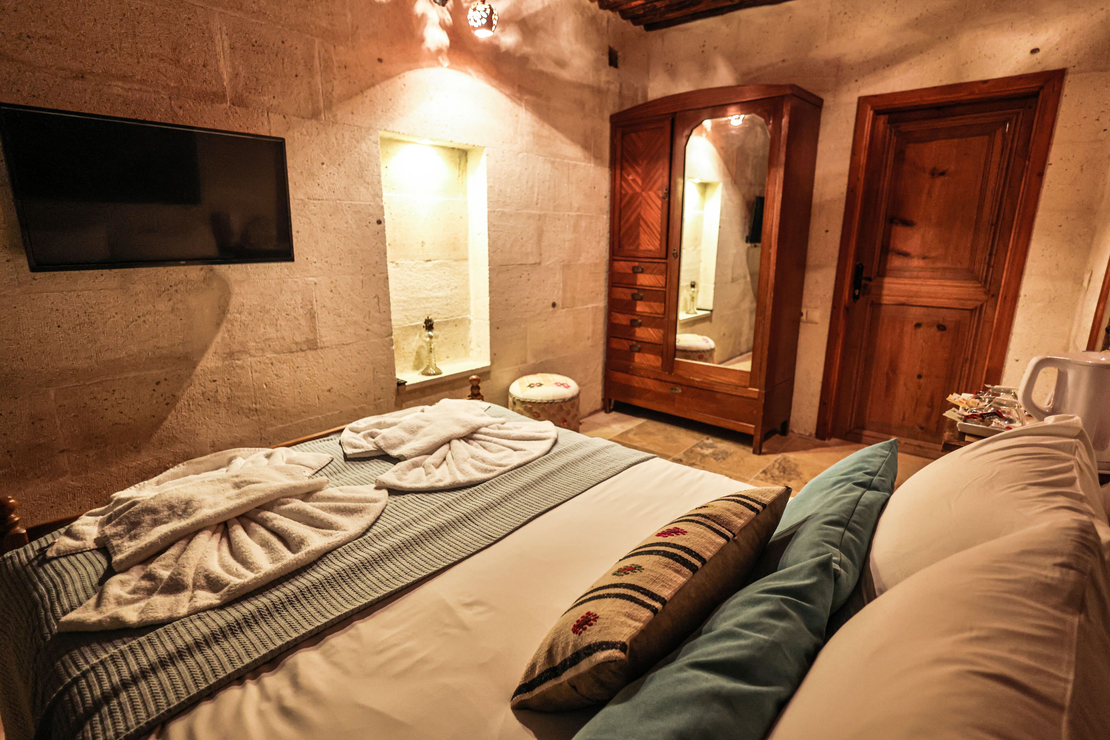 A bedroom with a bed, a wooden wardrobe, a mirror, and a television mounted on the wall.