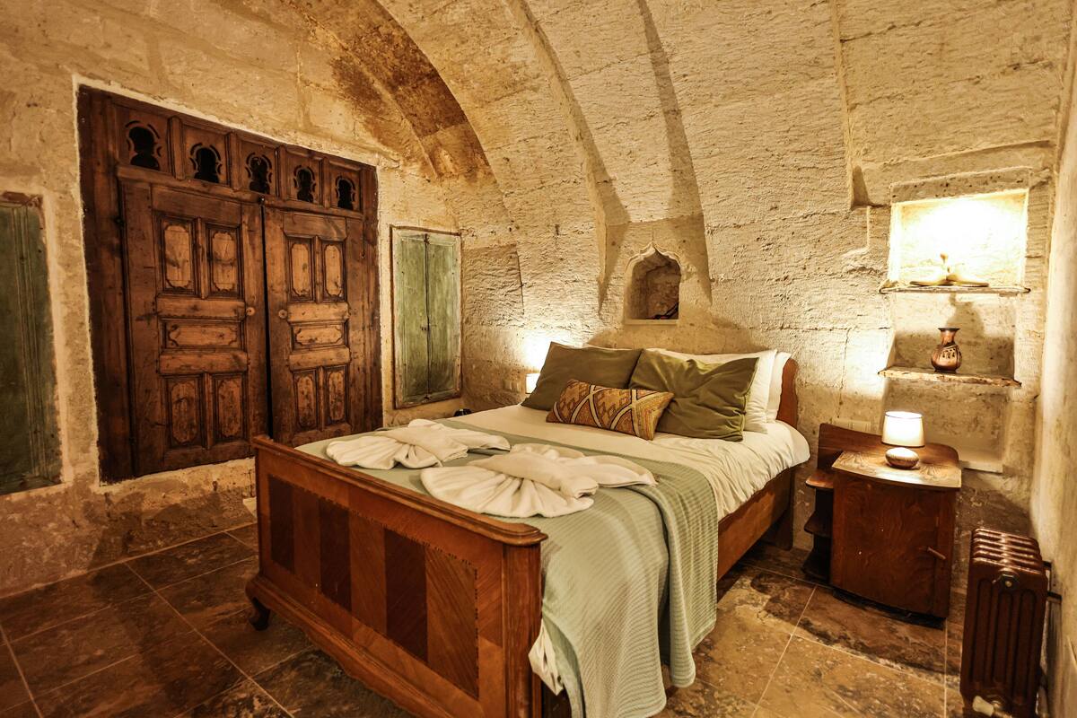 A rustic bedroom with a wooden bed, a stone wall, and a small bedside table with a lamp.