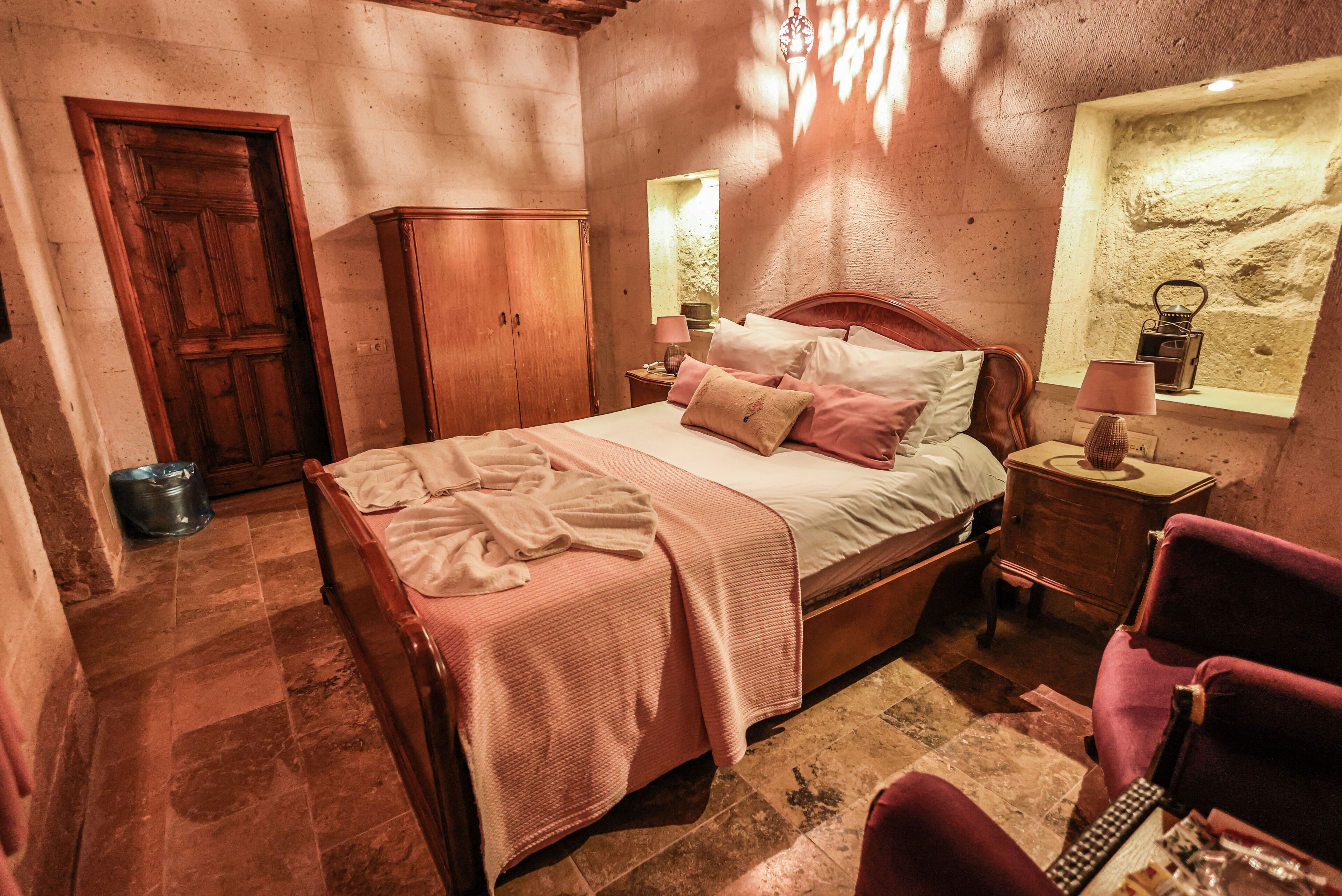 A bedroom with a wooden bed, a wardrobe, a nightstand with a lamp, and a stone wall.