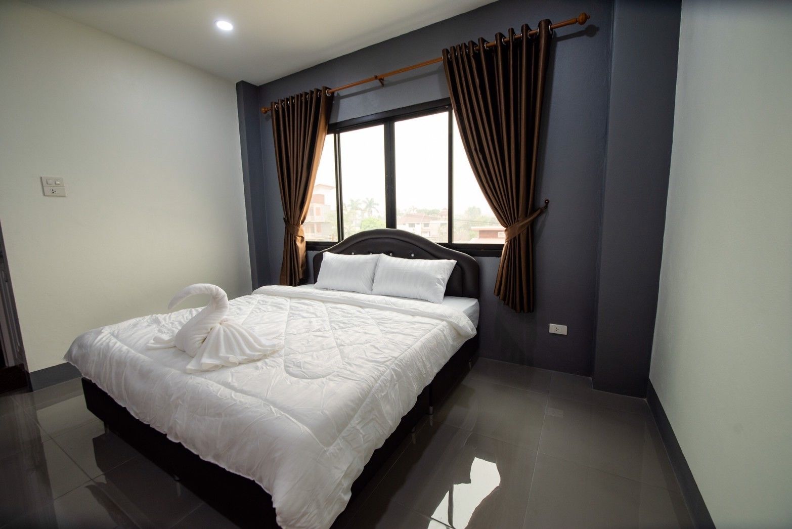 Una cama bien hecha en una habitación de hotel con un ventanal grande, paredes oscuras y cabecero de madera.
