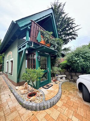 Un bâtiment pittoresque, doté d’un toit végétalisé, d’un petit porche, d’une plante en pot et d’un chemin de pierres menant à une voiture blanche.