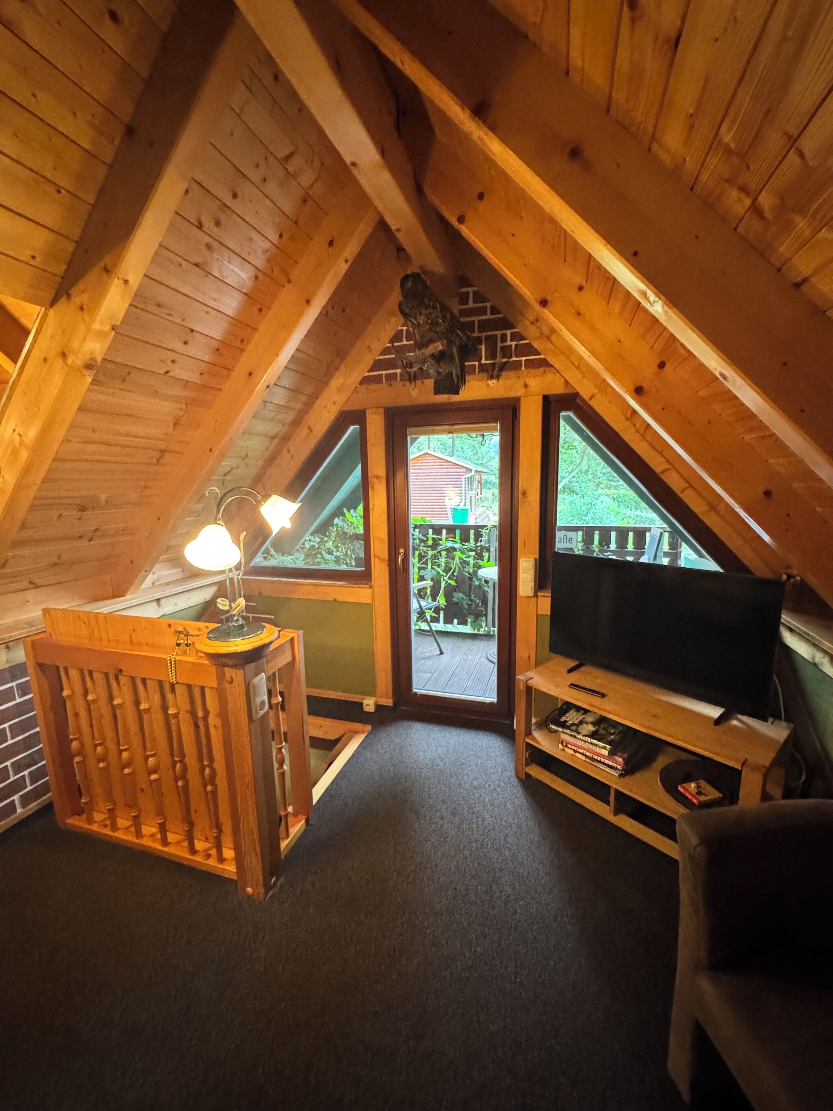 A cozy attic room with a wooden ceiling, a television, a small table with a lamp, and a door leading outside.