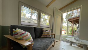 A modern living room with a grey sofa, colorful pillows, a wooden bench, and a glass table. Large windows provide views of the outdoors. - Vacation home close to nature with fireplace and rowing boat on Lake Plau - House 26 (Plau am See OT Quetzin)