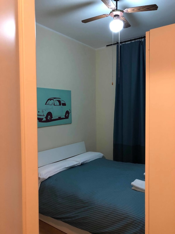 Una cama individual con una colcha azul, un almohadón blanco y una manta blanca. Hay un ventilador de techo con una lámpara arriba de la cama. Una cortina azul está parcialmente abierta, mostrando una habitación oscura detrás. Se ve una lámpara fijada en la pared en el lado izquierdo de la imagen.