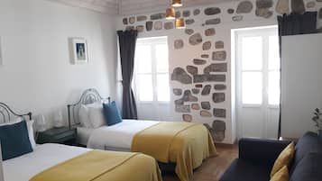 Ein Zimmer mit zwei Betten, einer Steinsäule, einem Fenster mit Vorhängen, einem Nachttisch mit Lampe und einem kleinen Bild an der Wand.