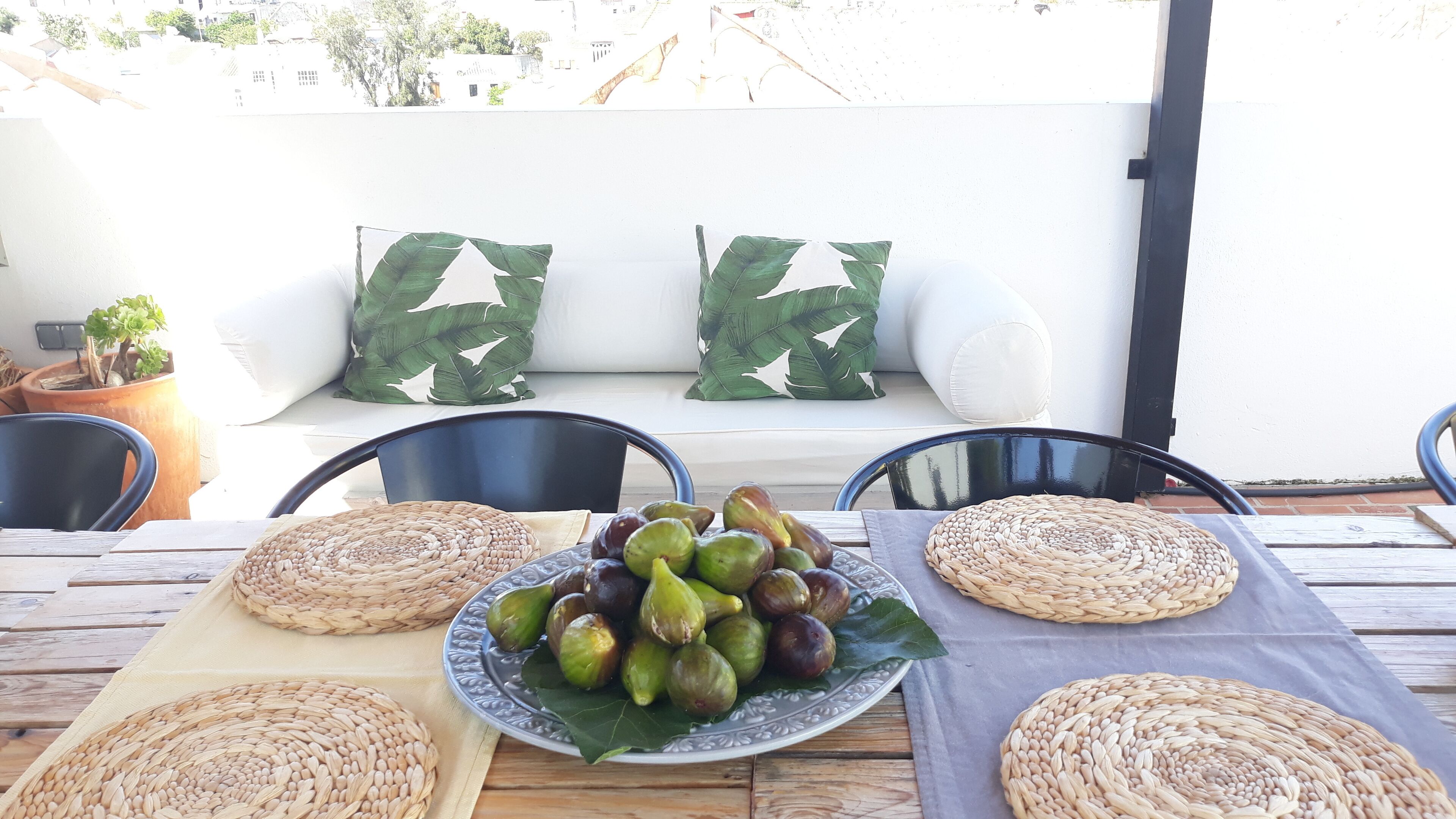 Une table recouverte de sets de table en osier, une assiette de figues et un canapé blanc agrémenté de coussins à motifs de feuilles.