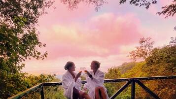 Two people in white robes sitting on a metal structure with a view of trees and a pink sky.