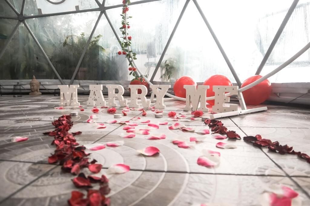A wedding setup with 'MARRY ME' letters, rose petals, and balloons.