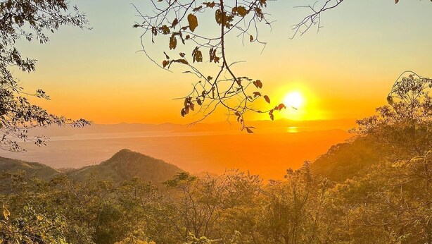A person sitting on a white blanket on a balcony overlooking a mountainous landscape during sunset. - El Mirador (Miramar)