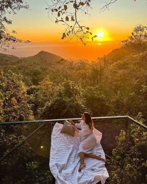 A person sitting on a white blanket on a balcony overlooking a mountainous landscape during sunset. - El Mirador (Miramar)