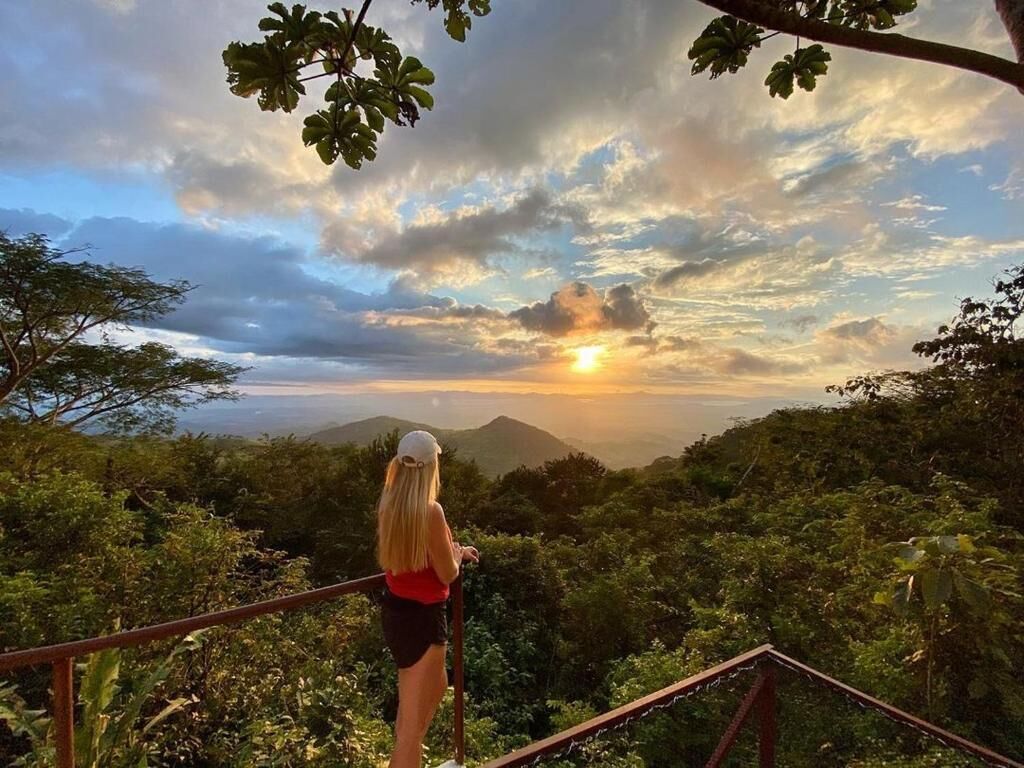 Uma pessoa está em uma varanda com vista para uma paisagem ao pôr do sol.