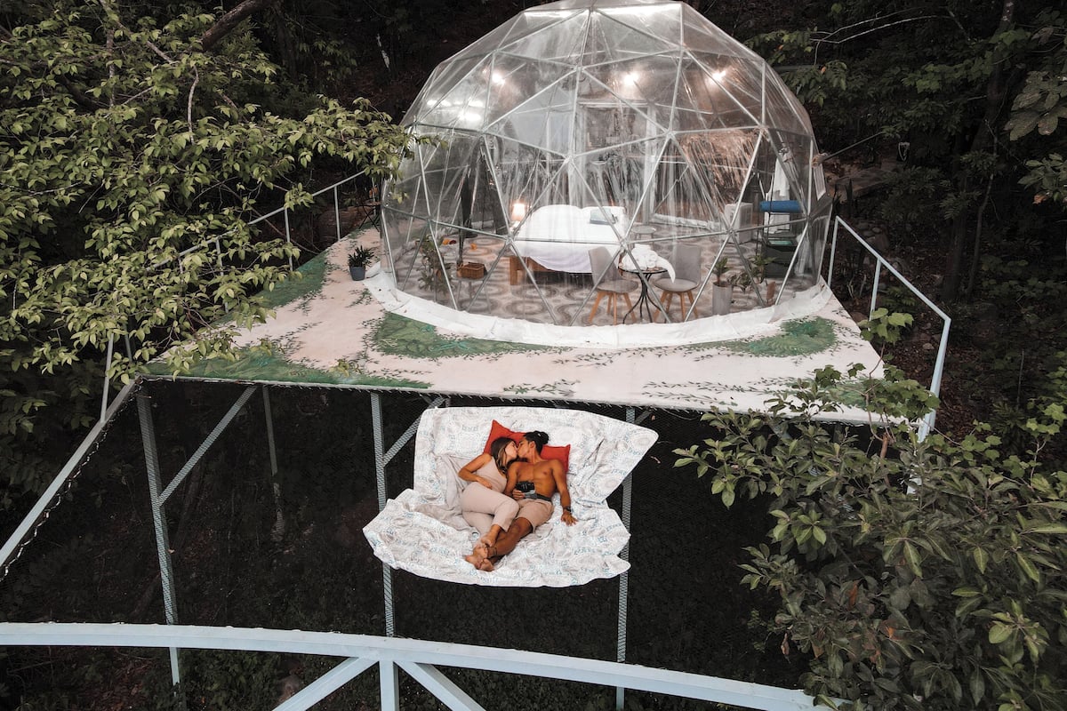 Estructura de cúpula geodésica transparente con una cama y zona de estar en su interior, rodeada de árboles.