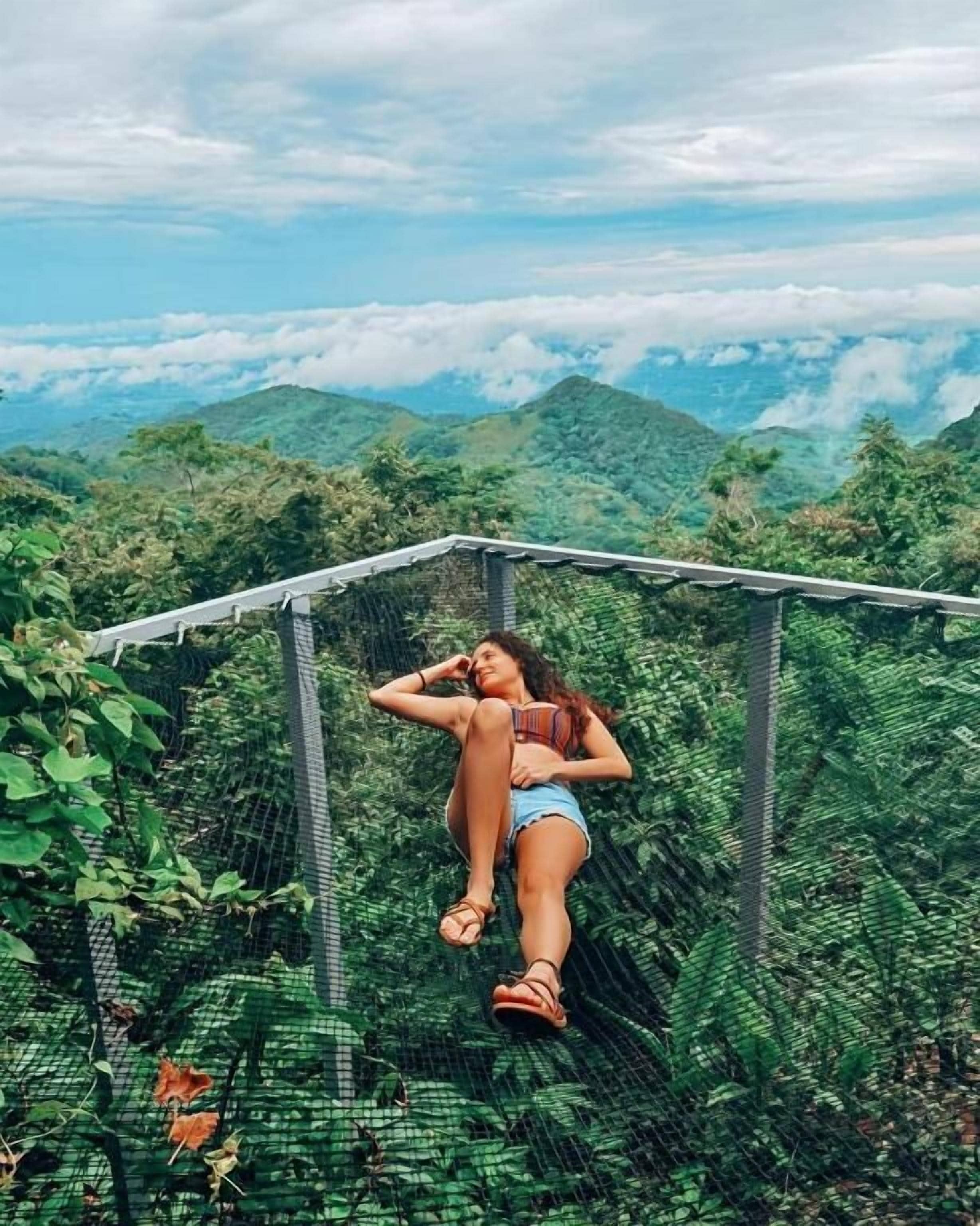 Una persona descansando en una hamaca en un área boscosa exuberante y verde, con vistas a montañas distantes.
