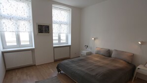 Ein Schlafzimmer mit einem großen Bett, zwei Nachttischen, einem Heizkörper und zwei mit Vorhängen versehenen Fenstern.