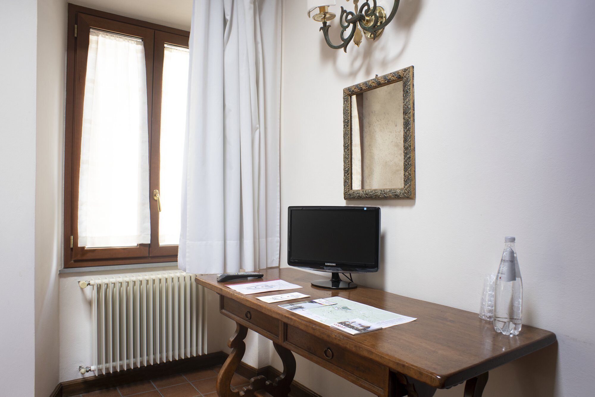Ein Zimmer mit einem Holztisch, einem schwarzen Monitor, einer Heizung, einem Fenster mit weißen Vorhängen, einem an der Wand befestigten Spiegel und einer Wasserflasche.