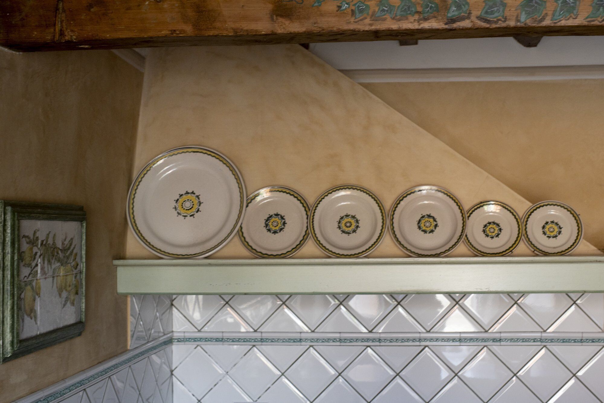 A row of six plates with a central design, arranged on a shelf above a tiled wall.