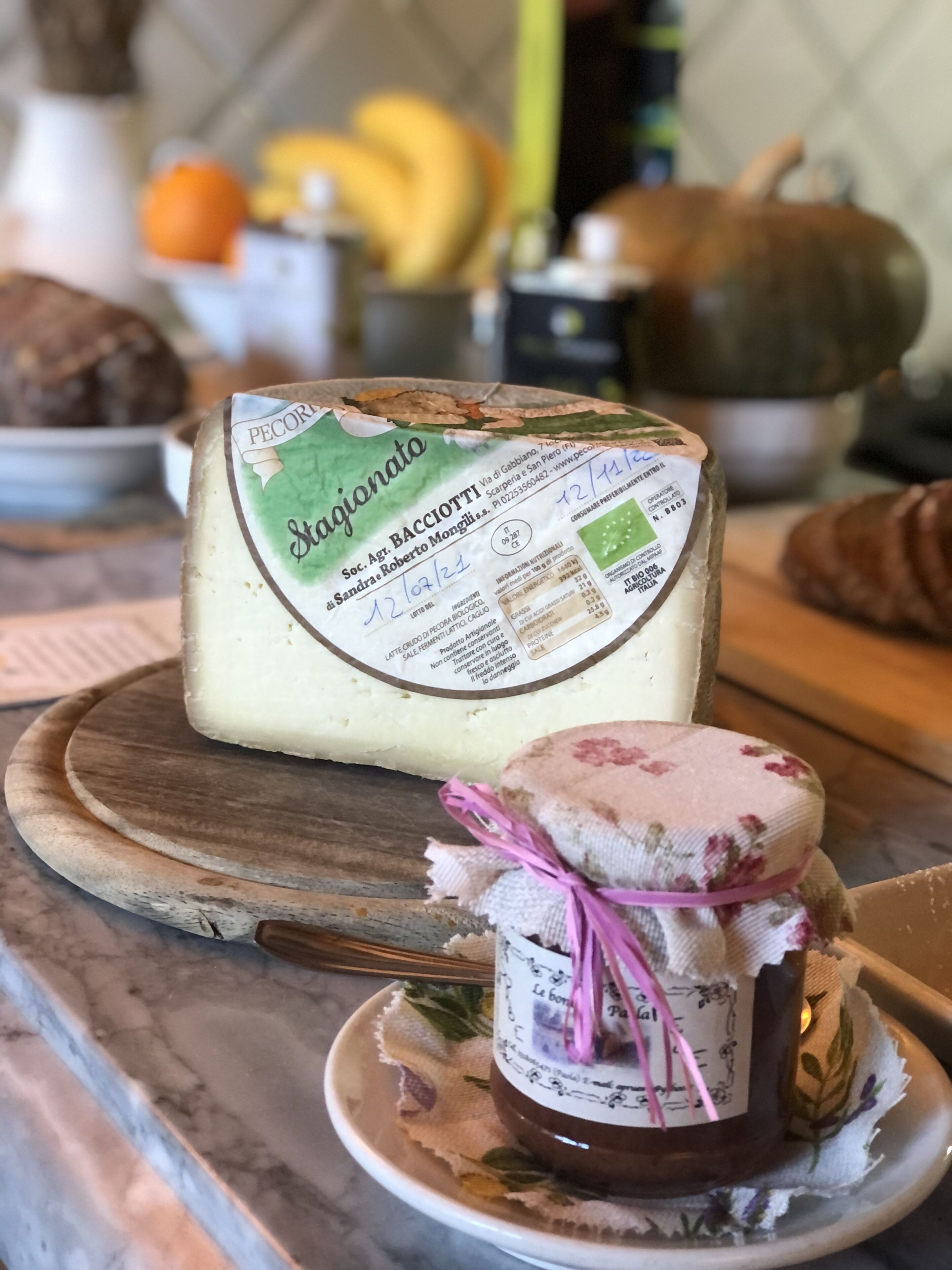 A wheel of Pecorino cheese on a wooden board, a jar of jam on a small plate, and a pumpkin in the background.