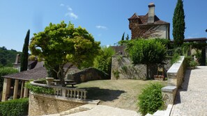 Un bâtiment en pierre, doté d’un toit en tuiles, entouré de verdure et bordé d’un chemin pavé.