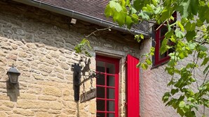 A stone building with a red door and window shutters, a sign reading "La Roserie," and a stone wall with a decorative vase.