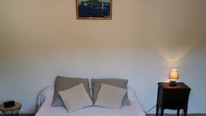 A neatly made bed with a white bedspread and two pillows, a small round table with a lamp, and a framed painting on the wall. - Charming villa in the heart of the maquis          (Bonifacio)