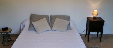 Una cama bien hecha con una colcha blanca y dos almohadas, una mesita redonda con una lámpara, y un cuadro enmarcado en la pared.
