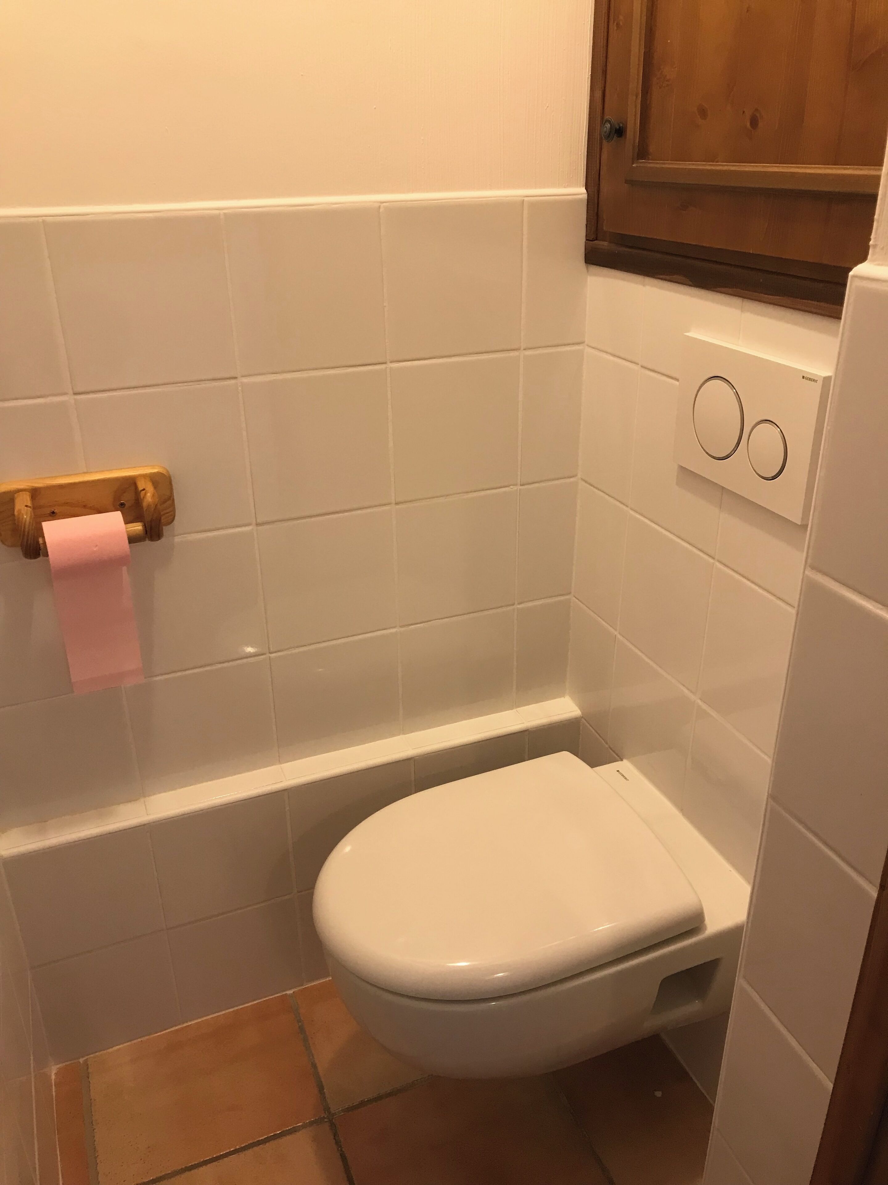 Un baño pequeño con azulejos blancos, un inodoro, un dispensador de papel higiénico de madera y un enchufe en la pared.