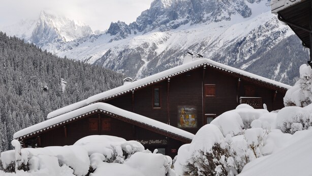 Un chalet en bois, doté d’un toit en pente, entouré d’arbres et de montagnes couverts de neige.