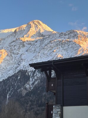 Ein Berg mit schneebedeckten Gipfeln und ein Holzbauten mit Steinmauer im Vordergrund.