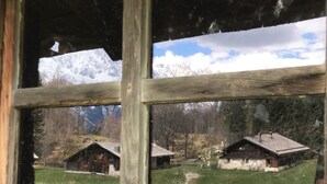 Un chalet rustique avec un cadre de fenêtre en bois, une cour verdoyante et une chaîne de montagnes en arrière-plan.