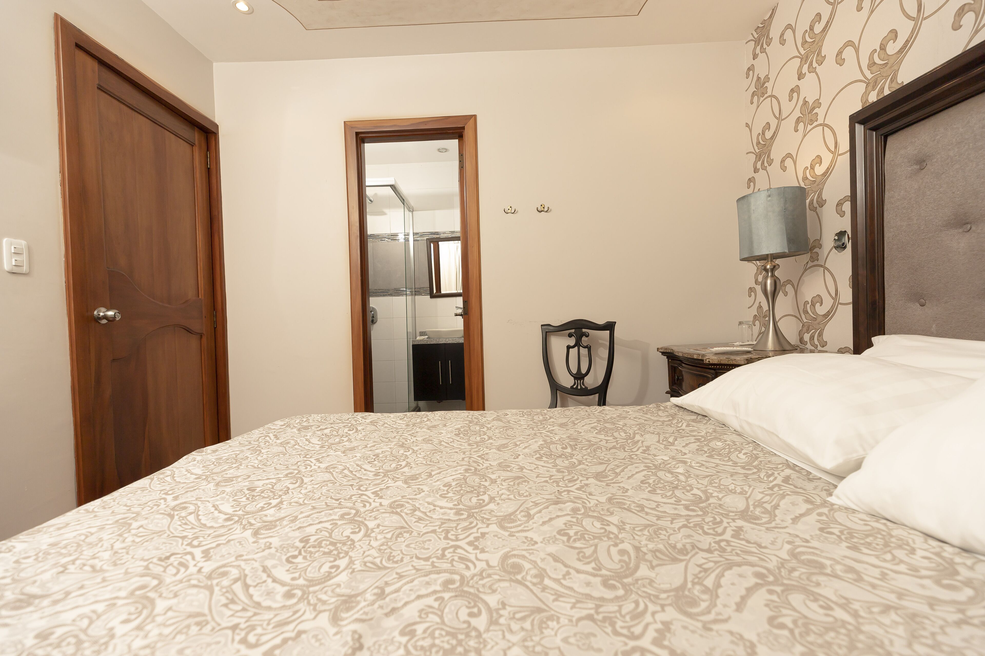 A bedroom with a patterned bedspread, a wooden door, a mirror, a bedside lamp, and a headboard.