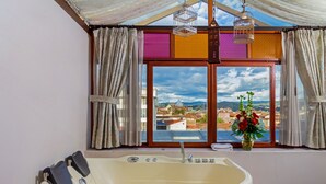 A bathroom with a bathtub, a window with curtains, and a view of buildings and hills. - Hotel Azul de la Plaza (Cuenca)