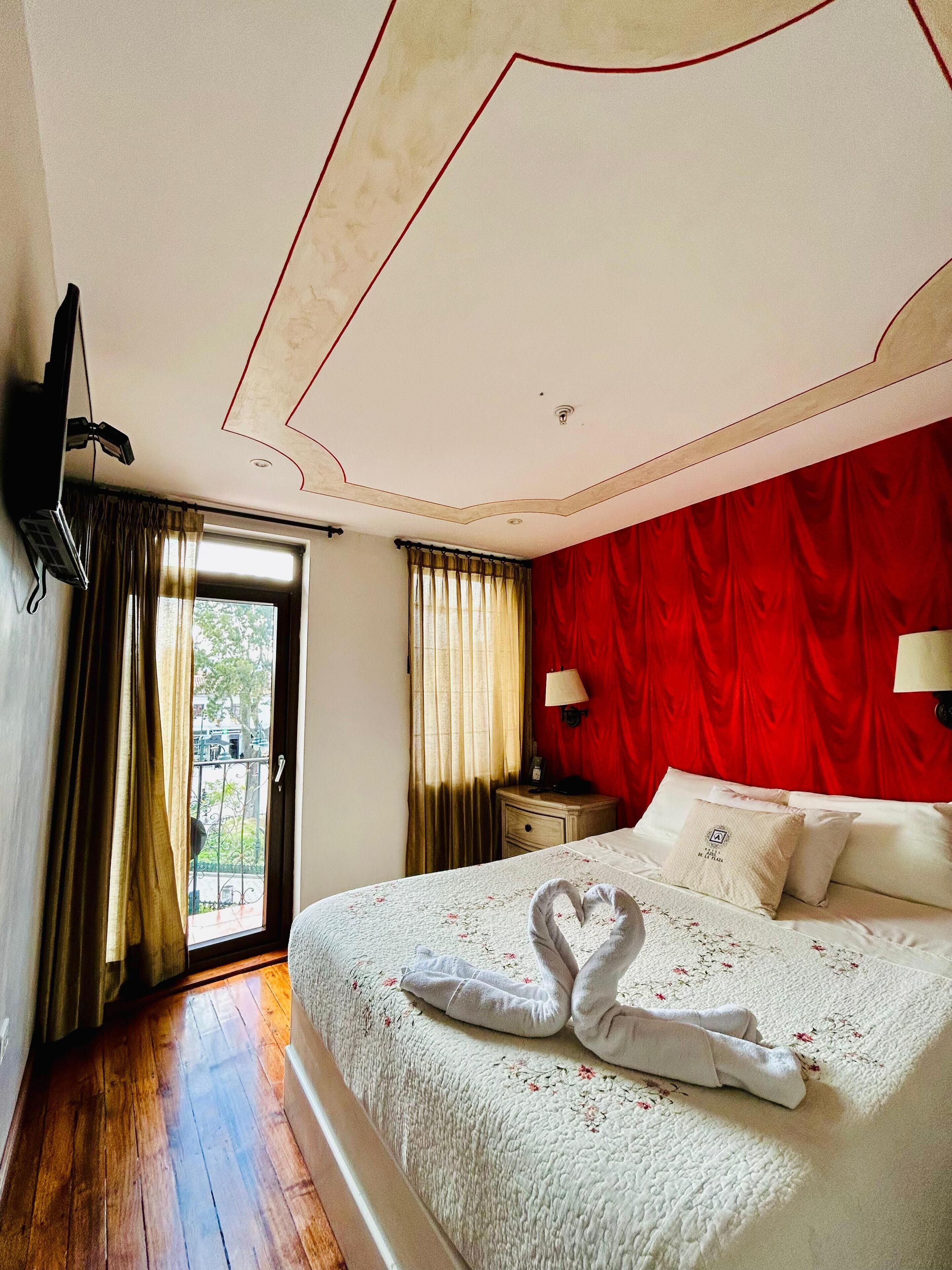 A hotel room with a bed, a TV mounted on the wall, a nightstand, a window with curtains, and a red wall.