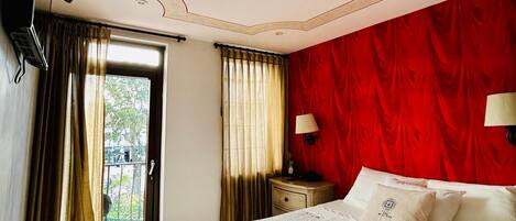 A hotel room with a bed, a TV mounted on the wall, a nightstand, a window with curtains, and a red wall.