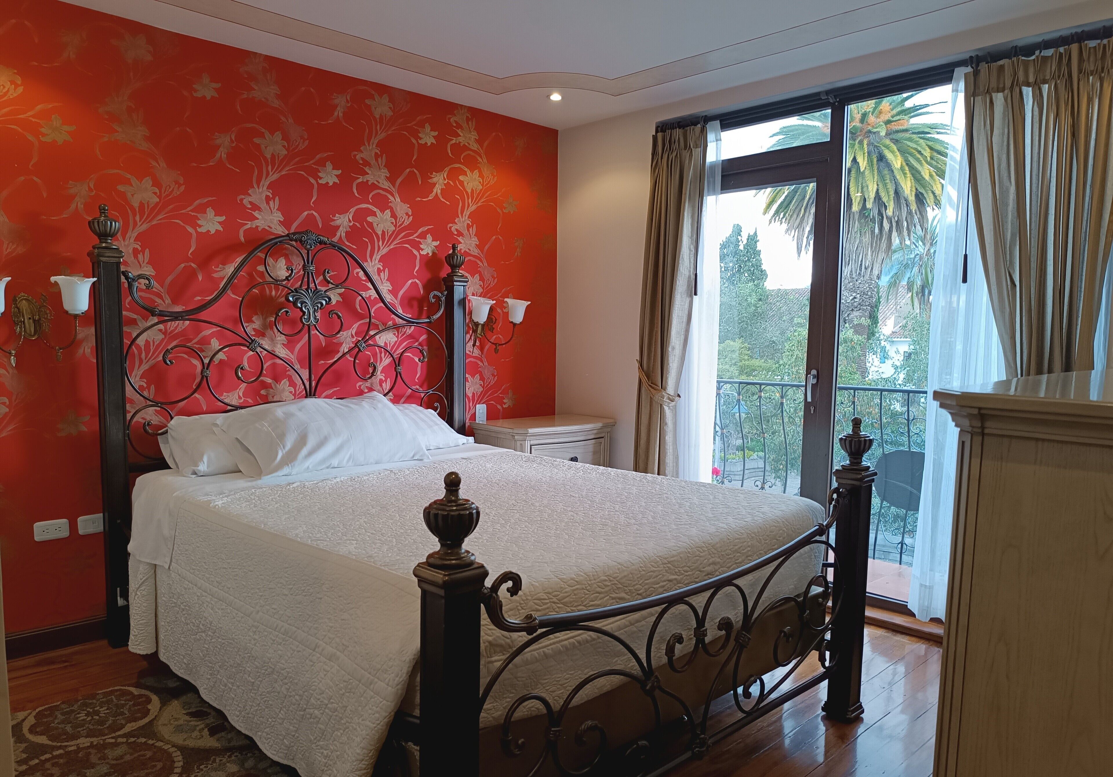 Un dormitorio con una cama grande, un papel tapiz rojo con motivos florales, una ventana con cortinas y un balcón con vistas a palmeras.