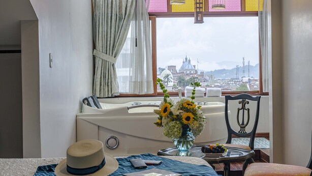 A hotel room with a large window offering a city view, a bathtub, a dining table with a vase of flowers, and a hat on the bed. - Hotel Azul de la Plaza (Cuenca)