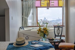 A hotel room with a large window offering a city view, a bathtub, a dining table with a vase of flowers, and a hat on the bed. - Hotel Azul de la Plaza (Cuenca)