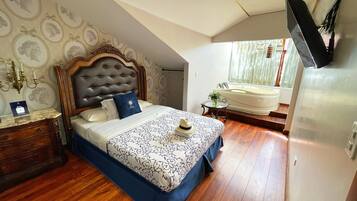 A bedroom with a wooden headboard, a bed with a patterned bedspread, a bedside table with a lamp, and a view of a bathroom with a bathtub.