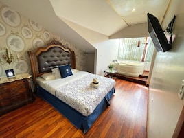A bedroom with a wooden headboard, a bed with a patterned bedspread, a bedside table with a lamp, and a view of a bathroom with a bathtub.