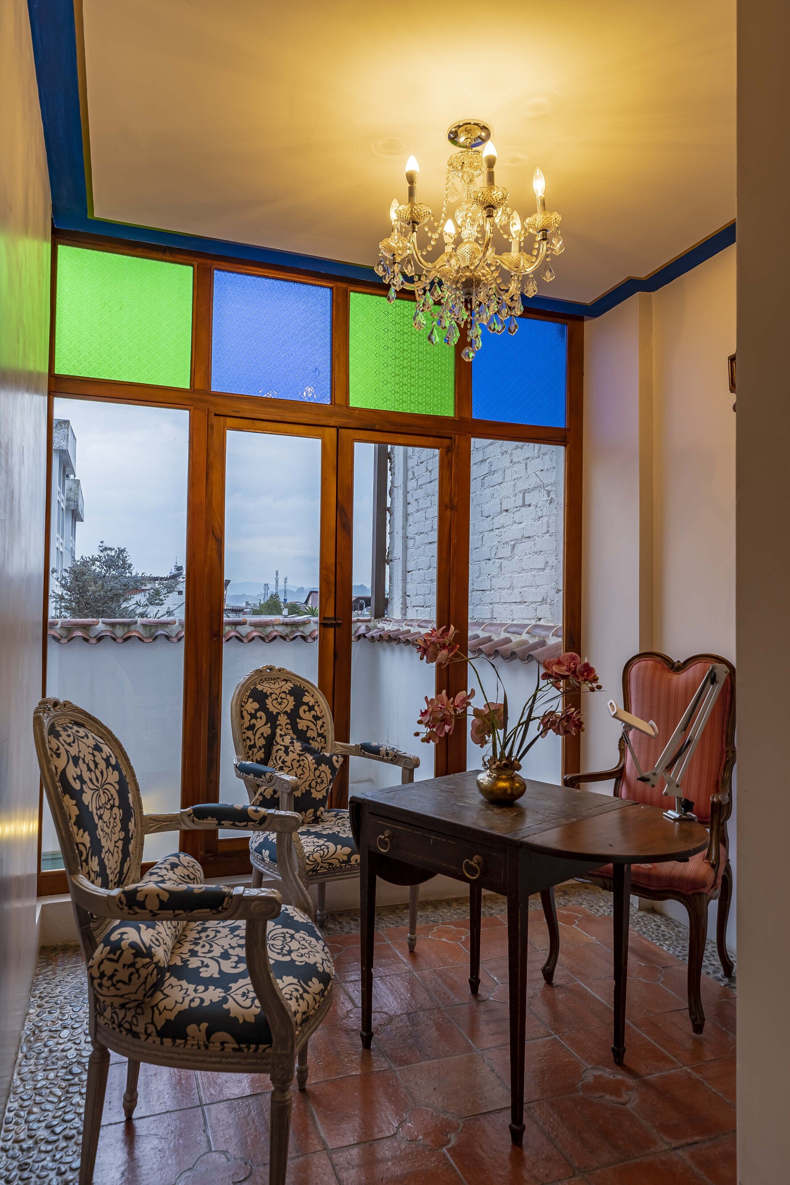 A dining area with a chandelier, patterned chairs, a wooden table, and large windows with a view of a building and trees.