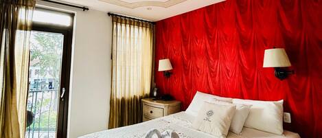 A hotel room with a large bed, red curtains, a wooden floor, and a view through a glass door.