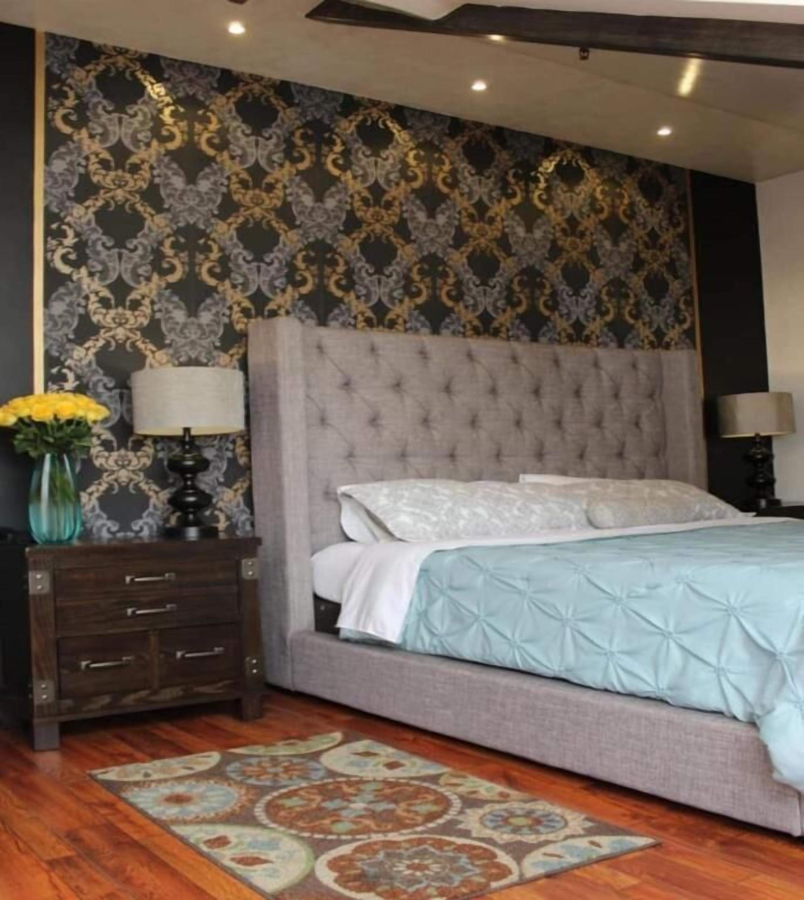 A bedroom with a tufted headboard, a patterned wall, a wooden nightstand, a lamp, a bed with a light blue bedspread, and a decorative rug.