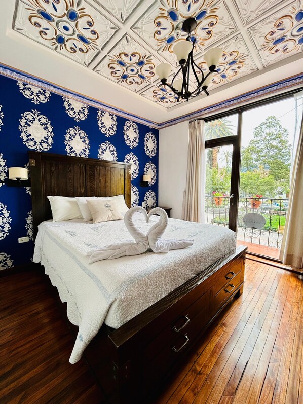 A bedroom with a large bed, a wooden headboard, a chandelier, and a balcony with a view of greenery. - Hotel Azul de la Plaza (Cuenca)