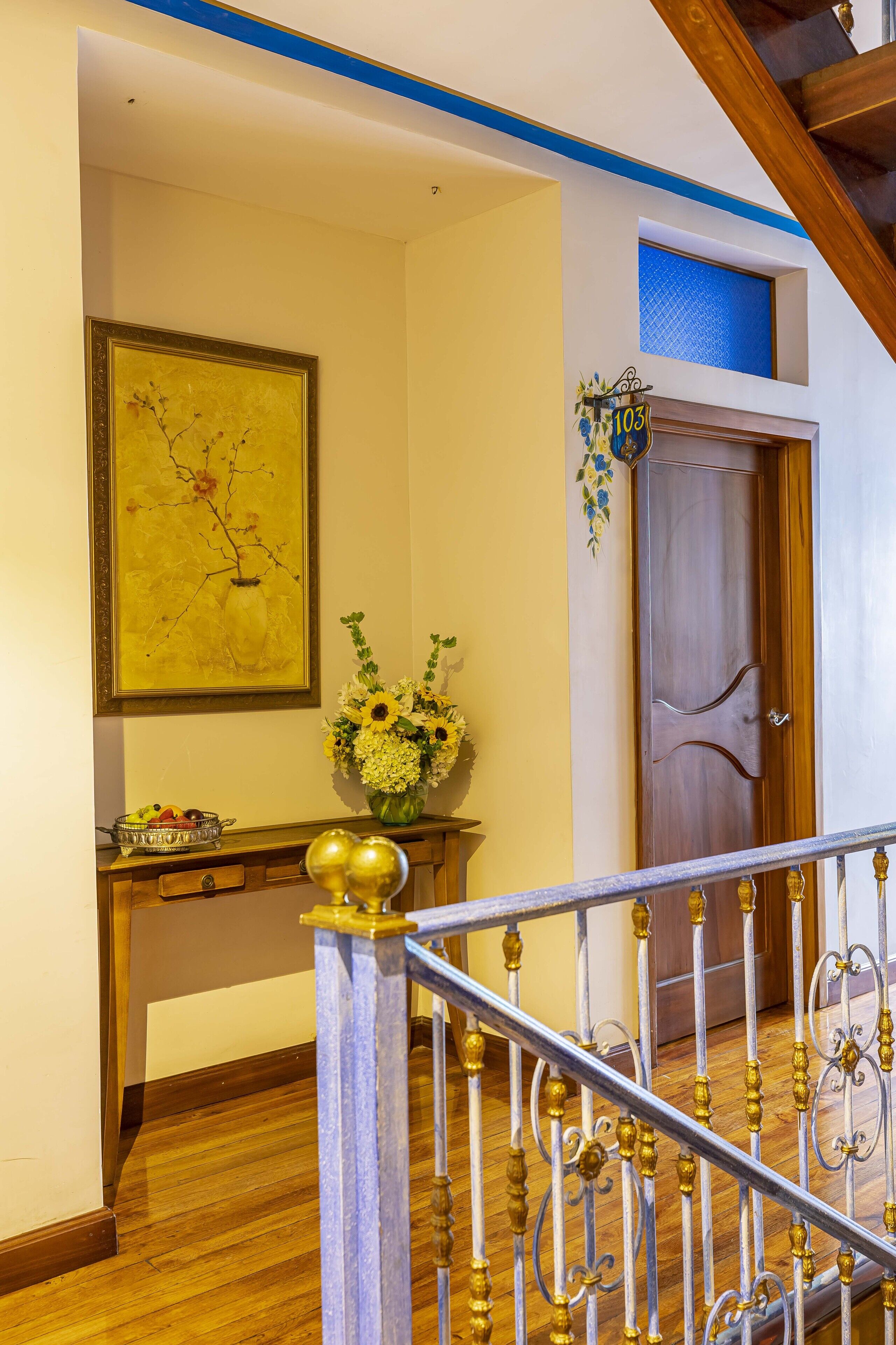 A wooden staircase with ornate metal railings, a wooden door with a number sign, a wall-mounted picture, and a vase with flowers.