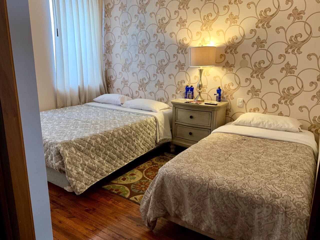 A hotel room with two single beds, a nightstand with a lamp, and a patterned wallpaper.