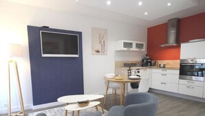 Una cocina moderna con armarios blancos, una pared de acento roja y un horno empotrado. Hay una zona de comedor con una mesa redonda y sillas, y un televisor montado en la pared.