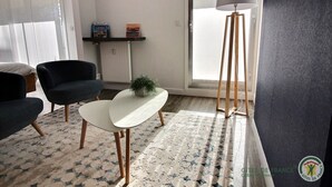 Una sala moderna con dos sillones negros, una mesa de centro blanca, una lámpara de pie y una planta pequeña. Se ve una estantería con libros y una ventana con vistas.