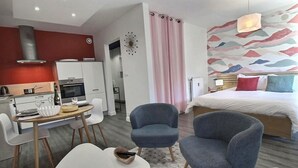 Una habitación de hotel moderna con una cama, dos sillones, un comedor con mesa y sillas, una kitchenette y un muro decorativo.