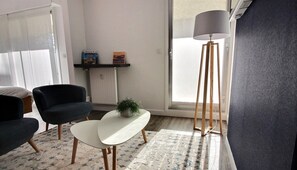 Ein modernes Wohnzimmer mit zwei schwarzen Sesseln, einem weißen Couchtisch, einer Stehlampe und einer kleinen Pflanze. Ein Bücherregal mit Büchern und ein Fenster mit Ausblick sind ebenfalls vorhanden.