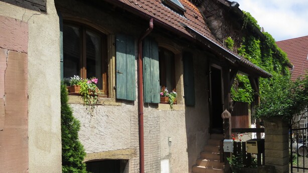 Un bâtiment traditionnel avec une façade en pierre, des volets en bois et un petit porche à l’avant, avec des marches menant à une porte.