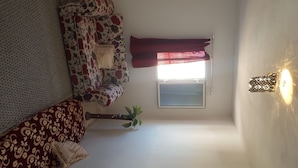 Habitació petita i acollidora amb un sofà estampat, una finestra amb cortines, una làmpada penjant a la paret i una planta en test.